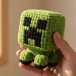 May include: A handmade green crochet Creeper from the video game Minecraft. The cube-shaped head features a black pixelated face. The Creeper has four small, rounded feet with black bottoms. The toy is held in a person's hand.