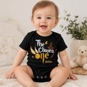 May include: A black short-sleeved baby bodysuit featuring the text The Chosen One in white and gold lettering, accompanied by a golden snitch, a brown sorting hat, a wand, and a lightning bolt graphic with the name Nathan printed below.