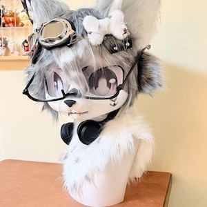 Puede incluir: Una cabeza de lobo antropomórfica de pelaje gris y blanco, con gafas de montura negra, gafas protectoras y auriculares. La cabeza tiene ojos grandes y de dibujos animados y un pequeño lazo blanco. La cabeza está montada sobre una forma blanca.