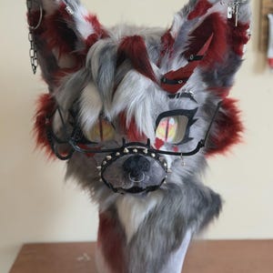 以下が含まれることがあります： グレー、白、赤の毛皮の狼の頭。マスクは大きな黄色の目、黒縁のメガネ、スパイク付きのマズルが付いています。アクセサリーには、チェーン、イヤリング、赤い羽が含まれます。頭は白い形にマウントされています。