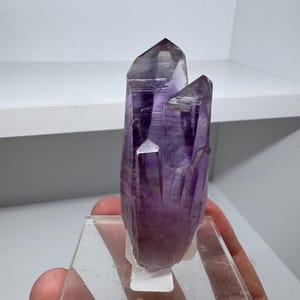 Puede incluir: Un grupo de cristales de amatista natural, que muestra un tono púrpura intenso con puntas translúcidas. El cristal está montado sobre un soporte acrílico transparente, resaltando su estructura geométrica y belleza natural. Un espécimen mineral.