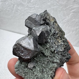 May include: A close-up of a mineral specimen featuring a cluster of metallic, dark gray, cubic crystals. The crystals are set against a matrix of dark green and black rock, showcasing natural geological formations.