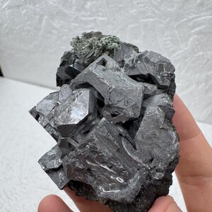 May include: A cluster of galena crystals, a metallic gray mineral, with a rough, textured surface. The crystals are cubic and intergrown, with a dark, almost black base. A small amount of green mineral is visible.