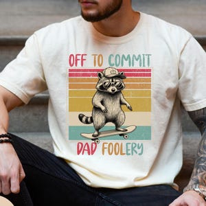 May include: Ivory-colored t-shirt featuring a raccoon wearing sunglasses and a cap, riding a skateboard. The shirt has the text "OFF TO COMMIT DAD FOOLERY" in a colorful retro design. The person in the image is wearing black jeans.
