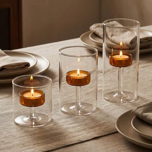 May include: Three clear glass candle holders with lit orange candles. The candles are set on a stem within the glass. The candle holders are on a table with plates and napkins.
