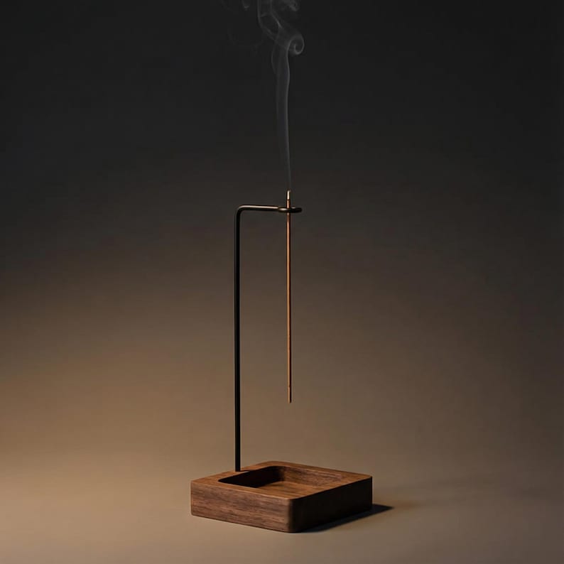 May include: A minimalist incense holder with a dark wood base and a black metal stand. A lit incense stick is suspended in the air, emitting a thin stream of smoke. The design is simple and elegant, suitable for home decor and aromatherapy.