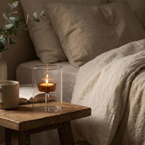 May include: A lit candle in a clear glass holder with a stem sits on a wooden table. The candle has an amber-coloured base and is enclosed in a tall, cylindrical glass. A book and a mug are also on the table. The background features a bed with neutral-toned bedding.