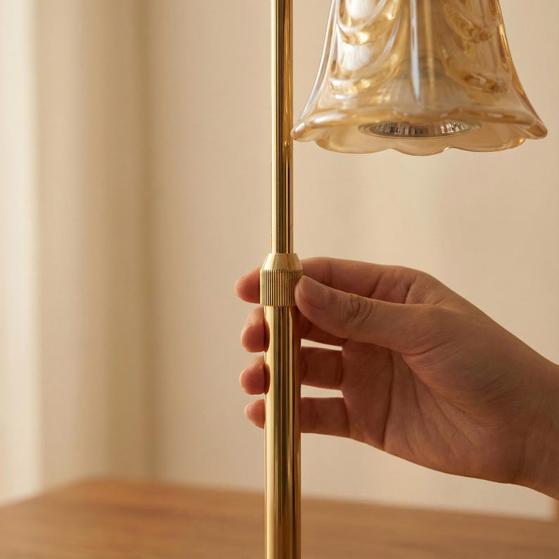 May include: A gold-colored lamp with a bell-shaped shade. The shade is a light amber color with a decorative pattern. The lamp's stem is a straight, gold-colored rod. A hand is holding the lamp.