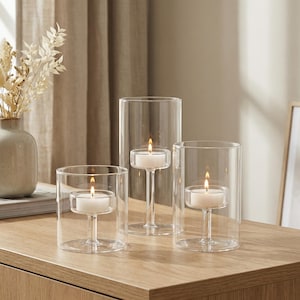 May include: Three clear glass candle holders with lit white candles. The candle holders are cylindrical, with a small platform to hold the candle. The candles are lit, casting a warm glow. The holders are on a wooden surface.