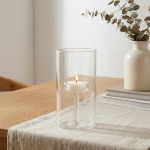 May include: A clear glass candle holder with a lit white tea light candle. The candle is suspended in the center of the glass cylinder. A small vase with greenery is in the background, along with a stack of books.