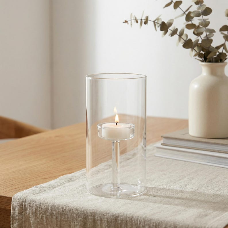 May include: A clear glass candle holder with a lit white tea light candle. The candle is suspended in the center of the glass cylinder. A small vase with greenery is in the background, along with a stack of books.