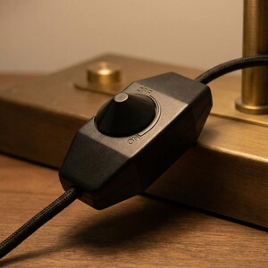 May include: Close-up of a black in-line lamp switch with a rounded toggle. The switch is labeled "OFF" and "ON" in white text. A black, braided power cord extends from the switch. The switch is set against a brass-colored lamp base.