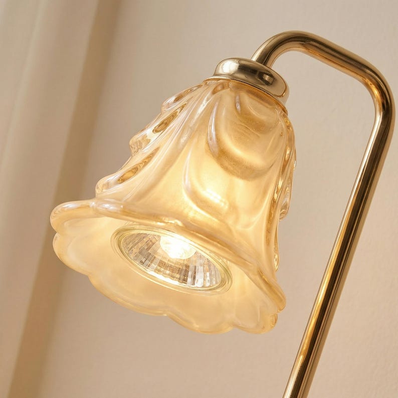 May include: A close-up of a decorative lamp with a gold-colored, curved metal frame. The lamp features a flower-shaped shade made of translucent, amber-colored glass. The light bulb is visible inside the shade.
