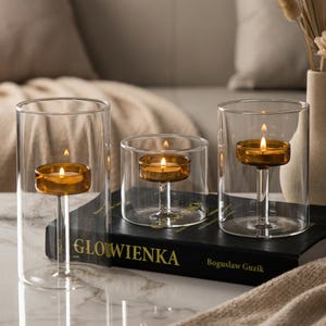 May include: Three glass candle holders with lit amber candles. The candles are set on a clear stem inside the glass. The candle holders are different sizes and are arranged on a book with the title "GLOWIENKA".