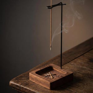 May include: A wooden incense holder with a starburst design carved into the base. A black metal stand holds a lit incense stick, with smoke rising. The holder sits on a wooden table, creating a warm, inviting atmosphere.