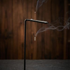 May include: A black metal incense holder with a minimalist design. Two incense sticks are lit, emitting wisps of white smoke. The holder has a horizontal bar to hold the incense and a vertical support. The background is a dark wooden surface.