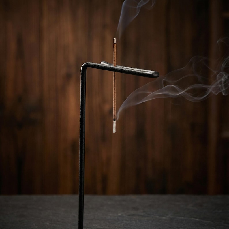 May include: A black metal incense holder with a minimalist design. Two incense sticks are lit, emitting wisps of white smoke. The holder has a horizontal bar to hold the incense and a vertical support. The background is a dark wooden surface.