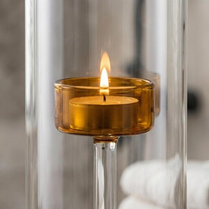 May include: A burning candle in a clear glass holder. The candle sits in a small, amber-tinted glass cup, with a bright flame. The holder has a long, clear stem and is enclosed in a larger glass cylinder.