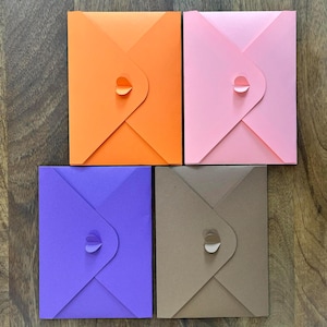 May include: Four colourful square envelopes in orange, pink, purple, and brown. Each envelope has a unique flap closure with a small, curved tab. The envelopes are arranged on a wooden surface.