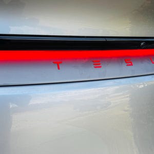 以下が含まれることがあります： シルバーのテスラ車のリアビューのクローズアップ。背面には赤い文字で「TESLA」の文字が表示されています。文字の上には明るい赤色のライトバーがあります。