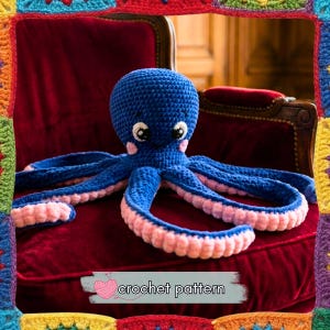 以下が含まれることがあります： 手作りの青いかぎ針編みのタコで、足にはピンクのアクセントが付いています。タコは大きくフレンドリーな目を持ち、頬にはピンクの斑点があります。画像には「crochet pattern」というテキストがバナーで表示されています。