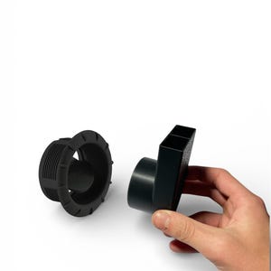 Puede incluir: Dos componentes de fontanería de plástico negro se muestran sobre un fondo blanco. Uno es una pieza cilíndrica con un borde exterior estriado y una abertura central. El otro es una pieza rectangular con un extremo circular.