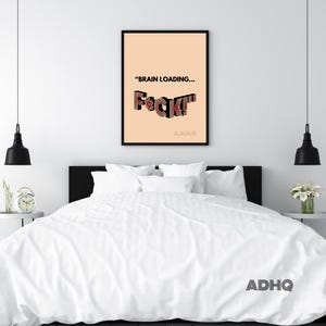 Puede incluir: Un cuadro enmarcado con el texto "BRAIN LOADING... F*CK!" en negro y rojo sobre un fondo melocotón. El cuadro está en un marco negro y cuelga sobre una cama con ropa de cama blanca. Las iniciales del artista, ADHQ, están en la parte inferior.