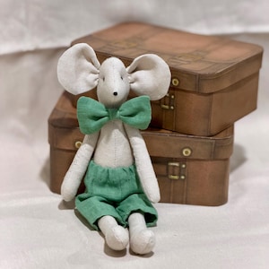 May include: A handmade stuffed mouse doll with large ears, a green bow tie, and green shorts. The doll is sitting in front of two brown vintage-style suitcases. The mouse is made of soft, light-colored fabric.