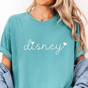 Puede incluir: Camiseta verde mar con la palabra "Disney" en escritura blanca. La palabra "Disney" tiene un coraz&oacute;n a cada lado. La camiseta es de corte holgado y tiene las mangas enrolladas.
