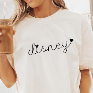 Puede incluir: Camiseta de color marfil con la palabra "Disney" en fuente cursiva, con detalles de corazones. La camiseta es de corte holgado. La modelo lleva calcetines blancos con rayas negras.