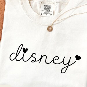 Puede incluir: Camiseta de color marfil con la palabra "disney" en escritura negra, con un icono de la cabeza de Mickey Mouse encima de la "d" y un coraz&oacute;n al final. La camiseta tiene cuello redondo y una etiqueta Comfort Colors.