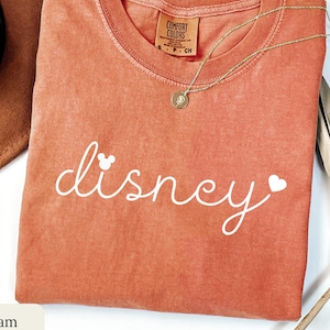 Puede incluir: Una camiseta Comfort Colors de color coral con la palabra "disney" en escritura blanca, con una cabeza de Mickey Mouse y un coraz&oacute;n. Un collar de oro con un colgante descansa sobre la camisa. Un sombrero marr&oacute;n y una hoja de palma seca tambi&eacute;n est&aacute;n en la imagen.