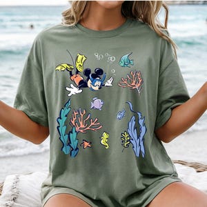 May include: Olive green t-shirt featuring a cartoon Mickey Mouse snorkeling underwater with colorful coral, fish, and bubbles. The design includes yellow flippers and a black snorkel mask. The shirt is a relaxed fit.