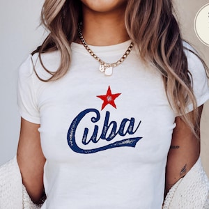 May include: White crop top with the word "Cuba" in blue script, topped with a red star. The shirt is a crew neck style. A gold chain necklace is worn.