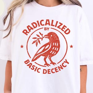 Peut inclure: T-shirt blanc avec un motif rouge. Le motif représente un oiseau tenant une branche dans un cercle, avec les mots "RADICALIZED BY BASIC DECENCY" disposés en cercle. Le t-shirt est de style décontracté.
