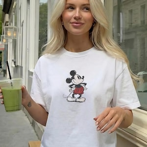 May include: White t-shirt with a Mickey Mouse graphic. The cartoon character is in black, red, and white. The shirt is a casual style, suitable for everyday wear.