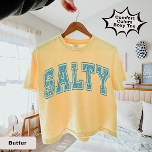 May include: A yellow t-shirt with the word "SALTY" in distressed blue lettering. The shirt is hanging on a wooden hanger. The text "Comfort Colors Boxy Tee" is in a black starburst shape.