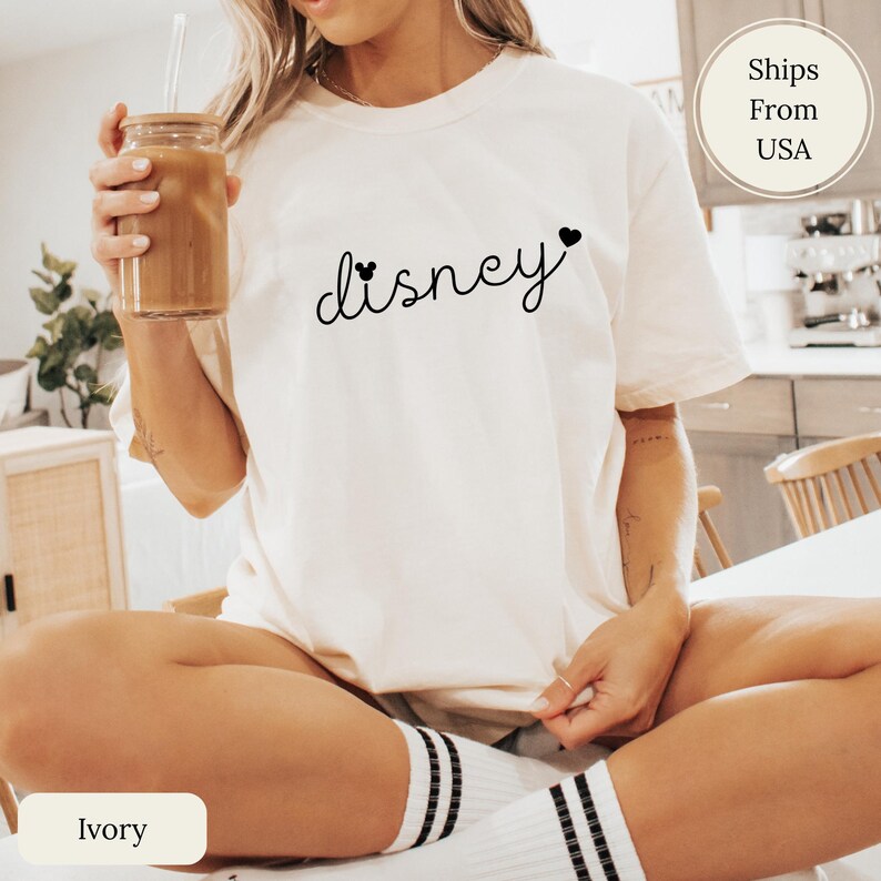 Puede incluir: Camiseta de color marfil con la palabra "Disney" en fuente cursiva, con detalles de corazones. La camiseta es de corte holgado. La modelo lleva calcetines blancos con rayas negras.