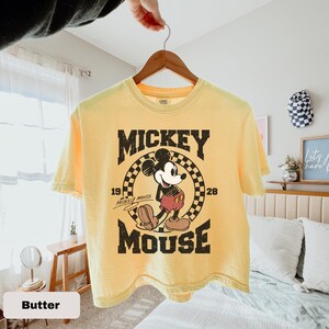 May include: Yellow t-shirt featuring a distressed graphic of Mickey Mouse. The design includes the text "Mickey Mouse" and the years "19" and "28". The shirt is hanging on a wooden hanger.
