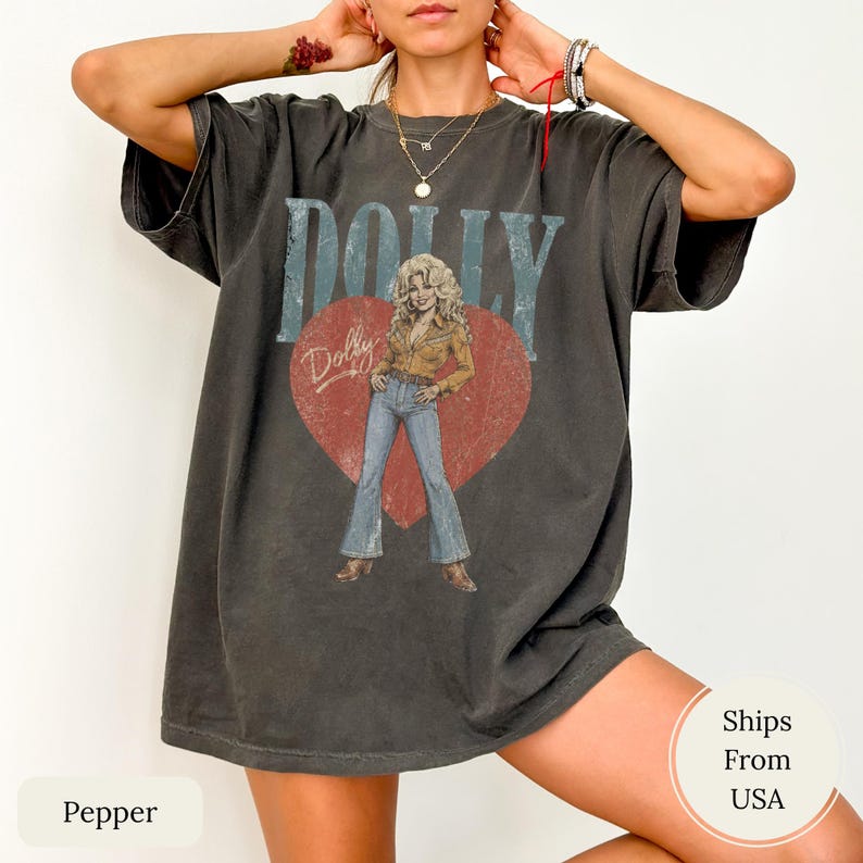 K&ouml;nnte beinhalten: Ein anthrazitfarbenes Kurzarm-T-Shirt mit einem gro&szlig;en roten Herz-Motiv und einer verwaschenen Illustration einer blonden Person in beigem Hemd und blauer Jeans. Das Wort DOLLY ist in gro&szlig;en, abgenutzten blauen Buchstaben &uuml;ber dem Herz gedruckt.