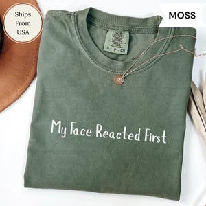 May include: A moss green t-shirt with the text "My Face Reacted First" in white. The shirt has a crew neck and short sleeves. A silver necklace with a round pendant is on the shirt.