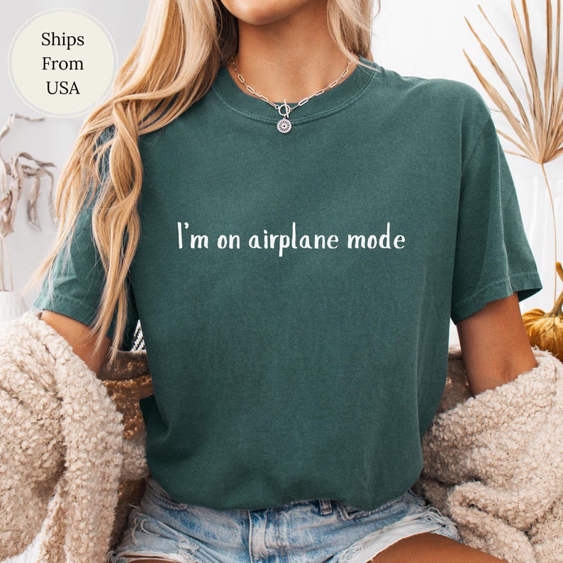 May include: A dark teal t-shirt with the phrase "I'm on airplane mode" in white lettering. The shirt has a crew neck and short sleeves. The image shows a person wearing the shirt.
