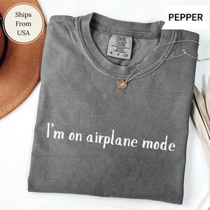 May include: A grey Comfort Colors t-shirt with the words "I'm on airplane mode" in white. A gold-coloured necklace with a pendant rests on the shirt. The shirt is folded and displayed on a white surface.