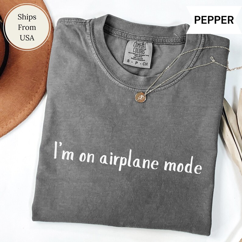 May include: A grey Comfort Colors t-shirt with the words "I'm on airplane mode" in white. A gold-coloured necklace with a pendant rests on the shirt. The shirt is folded and displayed on a white surface.
