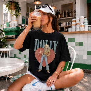 K&ouml;nnte beinhalten: Ein schwarzes Kurzarm-T-Shirt mit einem Grafikdruck einer Frau mit blonden Haaren, die ein beiges Hemd und eine blaue Jeans tr&auml;gt. Das Design enth&auml;lt ein gro&szlig;es rotes Herz, das Wort DOLLY in gro&szlig;en blauen Buchstaben und den Namen Dolly.