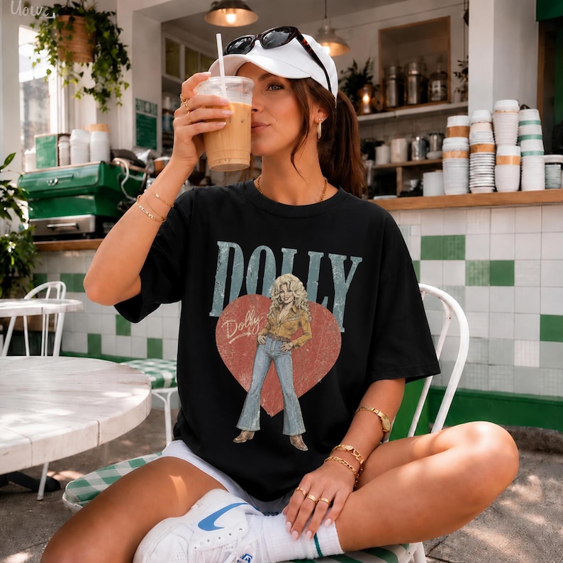 K&ouml;nnte beinhalten: Ein schwarzes Kurzarm-T-Shirt mit einem Grafikdruck einer Frau mit blonden Haaren, die ein beiges Hemd und eine blaue Jeans tr&auml;gt. Das Design enth&auml;lt ein gro&szlig;es rotes Herz, das Wort DOLLY in gro&szlig;en blauen Buchstaben und den Namen Dolly.