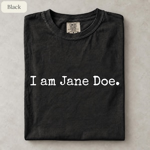 May include: A black short-sleeved t-shirt with a crew neckline. The shirt features the white text "I am Jane Doe." printed across the chest in a typewriter-style font. A Comfort Colors brand label is visible on the inside of the collar.