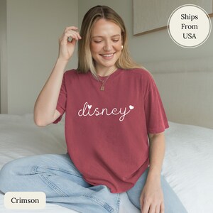 Puede incluir: Camiseta color carmes&iacute; con la palabra "disney" en escritura blanca, con detalles de corazones. La camiseta es de manga corta y cuello redondo. La modelo lleva vaqueros azules.