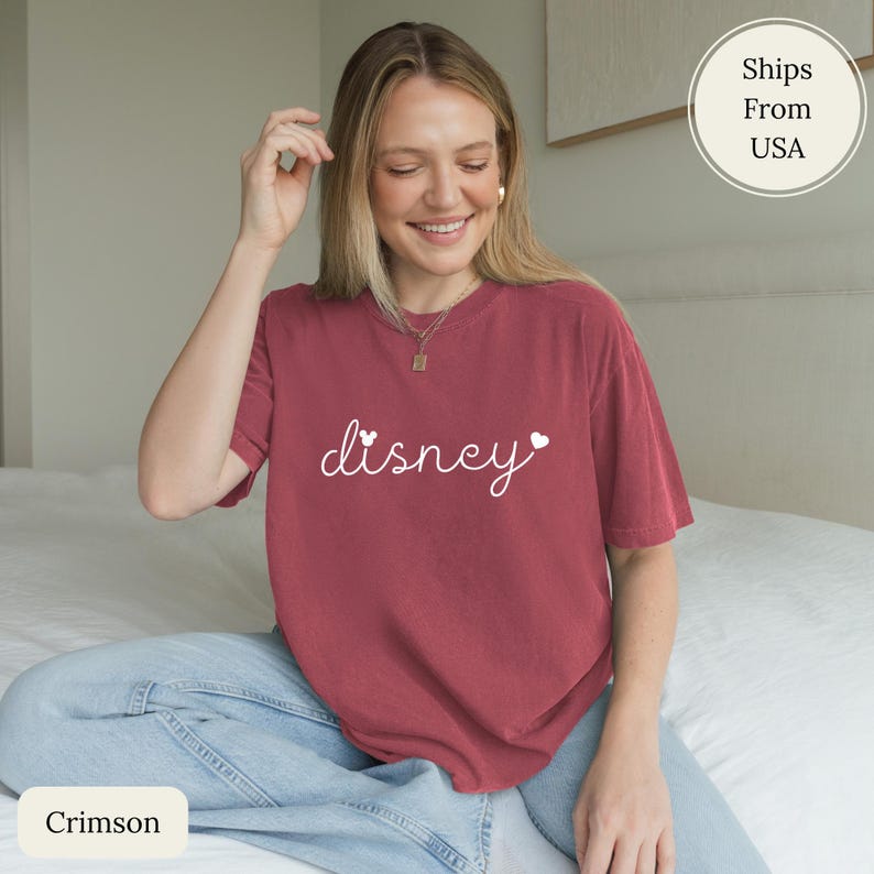 Puede incluir: Camiseta color carmes&iacute; con la palabra "disney" en escritura blanca, con detalles de corazones. La camiseta es de manga corta y cuello redondo. La modelo lleva vaqueros azules.