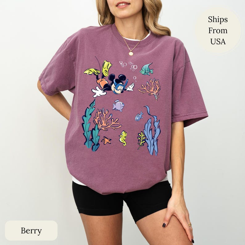 May include: Berry-colored t-shirt featuring a cartoon illustration of Mickey Mouse scuba diving underwater. The design includes colorful coral, fish, and bubbles. The shirt has a relaxed fit and is paired with black shorts.